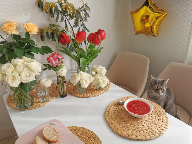 The cat sits at a table in a white kitchen with a plate of borscht against the background of bouquets of flowers