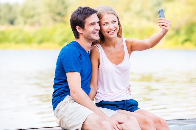 写真 人生の素晴らしい瞬間を捉えます。岸壁に座ってスマートフォンで自分撮りを作る美しい若い愛情のあるカップル