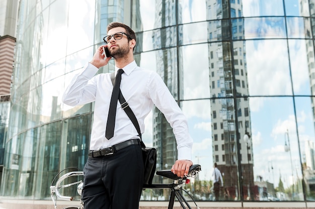 Catching up on the way to work. Low angle view of handsome young businessman talking on the mobile phone while leaning at the bicycle