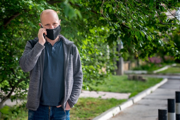 L'uomo adulto caucasico nella maschera protettiva nera sta camminando lungo una strada vuota e sta parlando al telefono. comportamento sicuro durante la pandemia di quarantena e coronavirus.