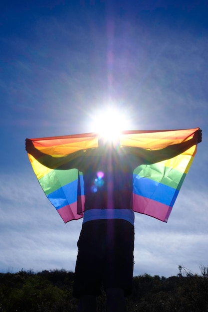 Caucasian adult in white t-shirt holds gay pride flag under blue sky.