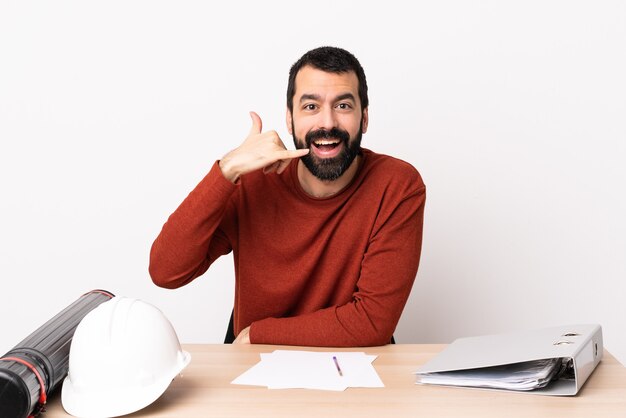 Caucasian architect man with beard in a table making phone gesture. Call me back sign.