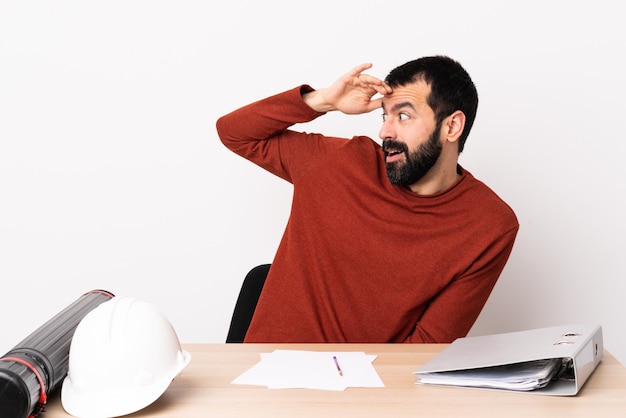 Uomo caucasico dell'architetto con la barba in una tabella con l'espressione di sorpresa mentre osservando lato