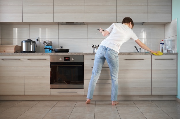 Caucasian bare feet girl weared in jeans and white t-shirt listening to music and cleaning a kitchen with yellow rag