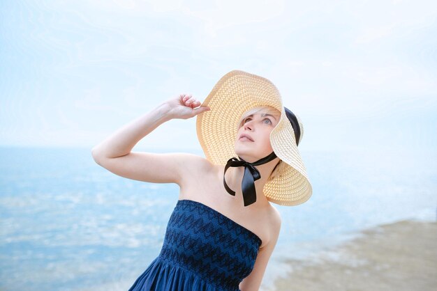 Caucasian beautiful stylish blonde sad woman in blue dress and in straw hat standing near the sea