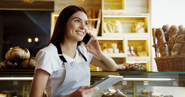 Caucasian beautiful woman baker or bread seller with a smile talking cheerfully on the phone while holding tablet device in hands and looking at its screen in the bakery shop. Indoor