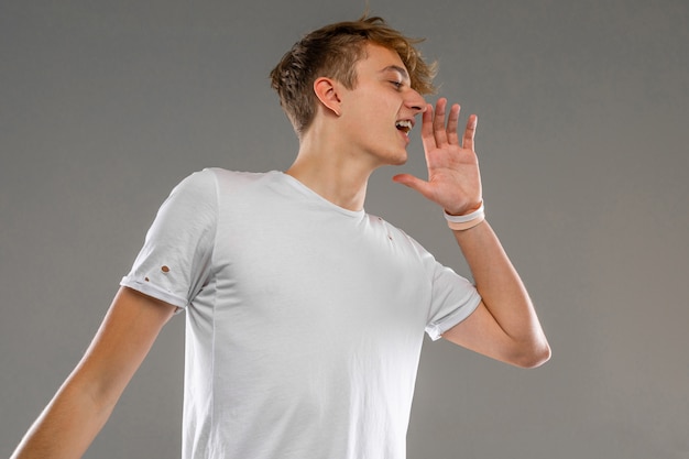 caucasian blond guy in a white t-shirt is calling someone holding his hand near his mouth on a gray wall