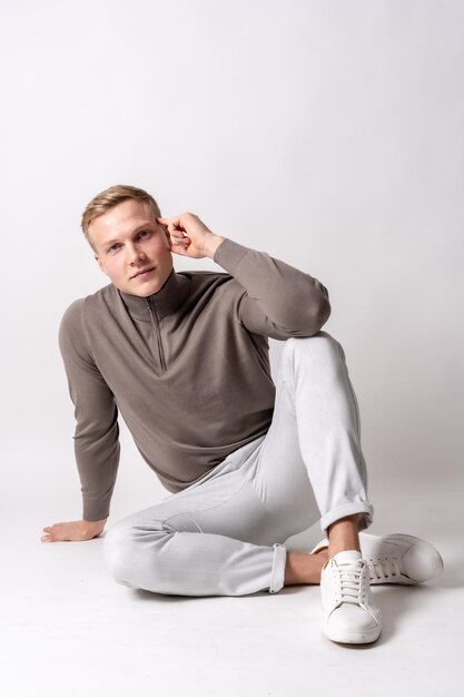 Caucasian blond model with a brown sweater on a white background sitting on the floor with a seductive look