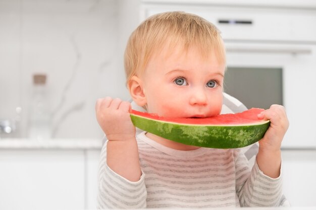 Caucasian blonde baby 1 year old have lunch at kitchene ating red water melon healthy food