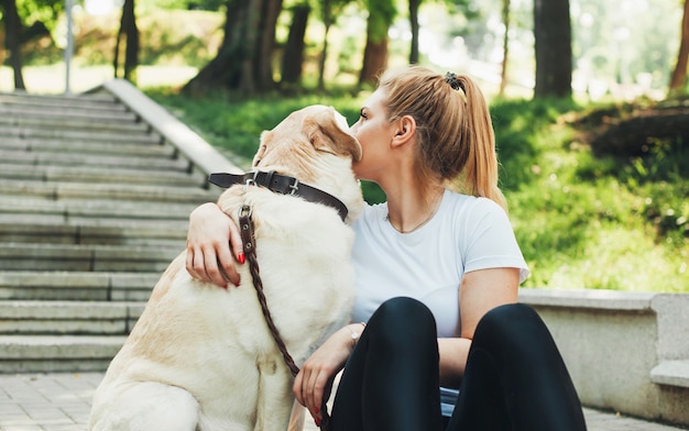 Caucasian blonde woman is embracing her labrador during a rest in park on the stairs