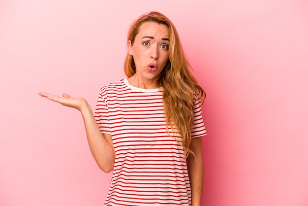 Caucasian blonde woman isolated on pink background impressed holding copy space on palm.