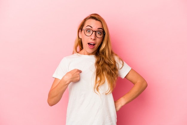 Caucasian blonde woman isolated on pink background surprised pointing with finger, smiling broadly.