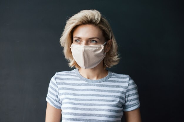 Caucasian blonde woman looking somewhere posing on a gray studio wall with a medical mask on face