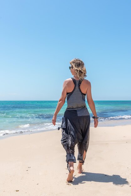 Caucasian blonde woman walking along a golden sandy beach in casual clothes on a sunny day