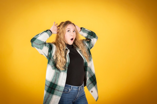 Donna bionda caucasica che indossa abiti casual, camicia a quadri verde, sorpresa su sfondo giallo