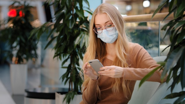 Caucasian blonde woman wearing medical mask in shopping center mall company looking at phone screen