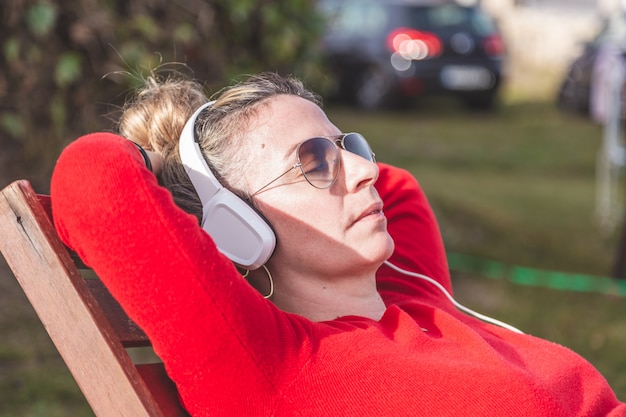 Caucasian blonde woman with red sweater sunglasses and  white headphones listening to music and relaxing outdoors.