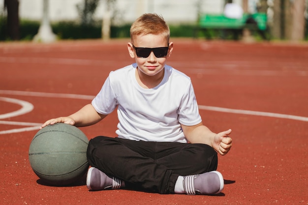 白人の男の子のブロンドは、スポーツグラウンドでバスケットボールのボールと一緒に座っています