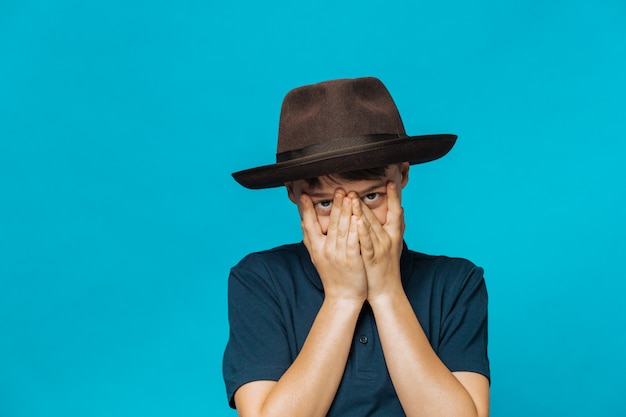 Foto ragazzo caucasico vestito con una maglietta blu scuro e un cappello con un bordo, si copre il viso con le mani in una timida espressione facciale e guarda attraverso le dita sorridendo