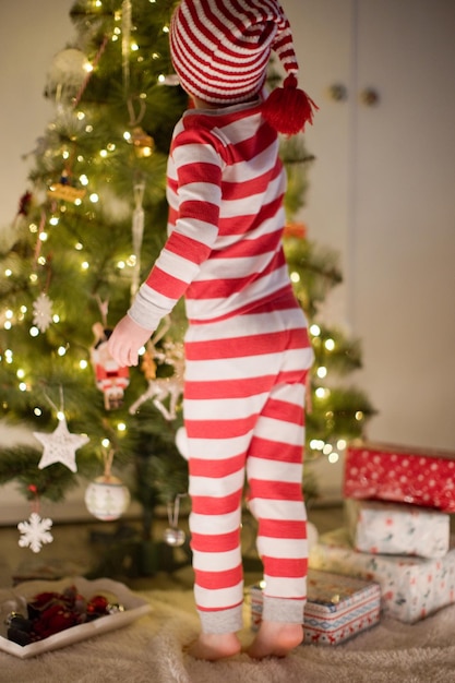 Caucasian boy in striped pajamas and hat standing near Christmas tree Cozy Christmas interior with decorations and lights