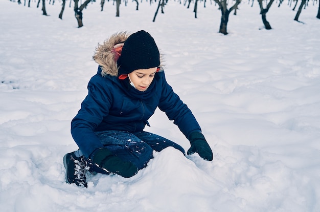 暖かくて帽子をかぶった白人の男の子が雪で遊んでいます。