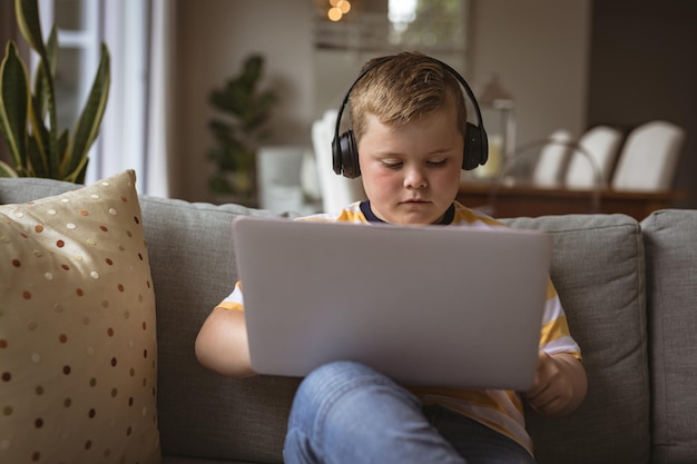 Caucasian boy wearing headphones using laptop sitting on the couch at home