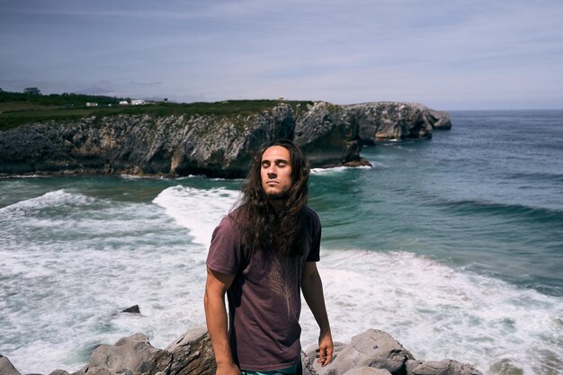 Photo caucasian boy with long loose disheveled hair with closed eyes tshirt on the rocks by the sea near