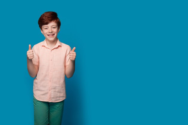 Caucasian boy with red hair is gesturing the like sign on a blue studio wall with free space advertising something