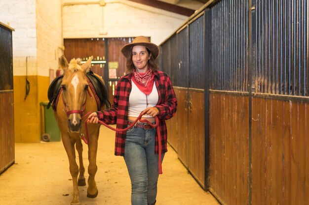 Caucasian brunette cowgirl woman strolling with a horse in a stable, wearing American cowboy hat, red plaid shirt and jeans