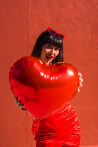 Caucasian brunette girl with heart balloons on Valentines day wearing sunglasses red background Very in love the day of celebration a balloon