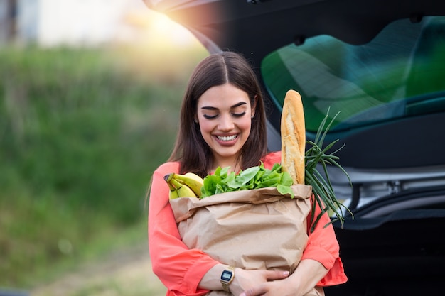 Caucasian brunette going holding paper bags with food products. Young woman putting package with groceries and vegetables into car trunk.