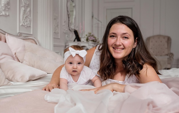 Caucasian brunette mom lies on the bed with her baby daughter and looks at the camera Happy motherhood