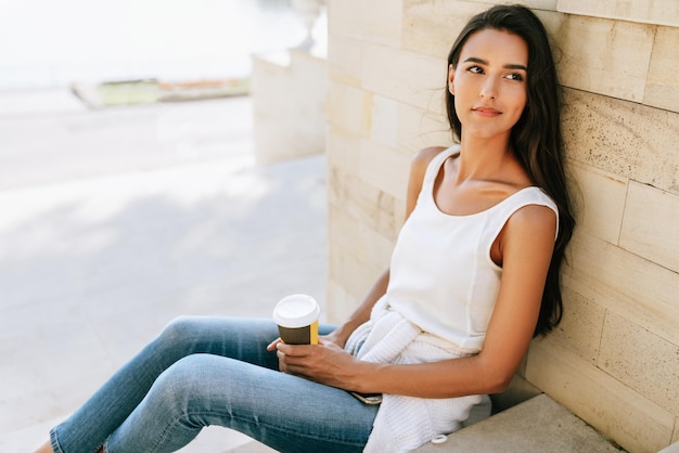 Caucasian brunette woman holding disposable cup of coffee\
sitting on the stairs in the city street pretty female takeaway\
coffee in hand pretty student drinking coffee outdoors in the\
morning