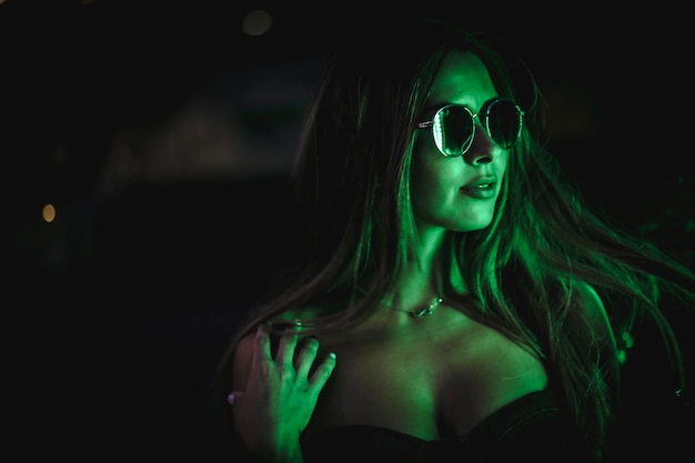 Donna caucasica del brunette con gli occhiali da sole illuminati con la luce verde dei led. fotografia urbana notturna