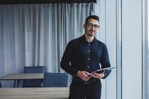 A Caucasian business man in glasses and a black shirt sits in the office and looks at sales growth charts A young successful businessman in classic clothes stands by the window in the office