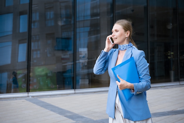 Caucasian business woman in a blue jacket and dress talking on the phone with a folder of papers in her hand against the wall of an office building