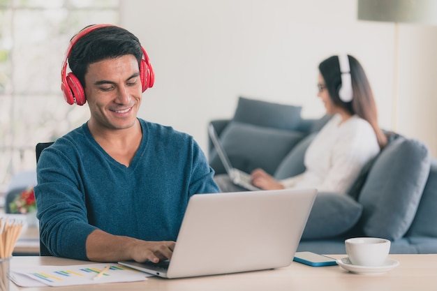 Caucasian businessman and Asian businesswoman wearing casual dress sitting together in living room and workin on laptop notebook computer white put on headphones. Idea for modern working at home.