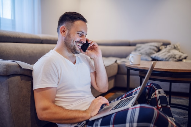 Uomo d'affari caucasico in pigiama seduto sul pavimento in salotto, usando il portatile e parlando al telefono intelligente al mattino.