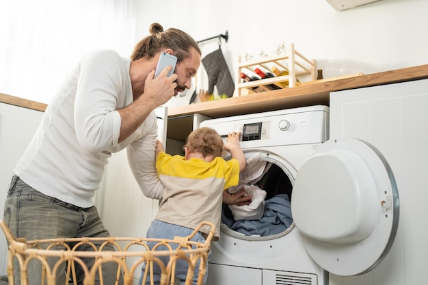 caucasian busy father doing housework with baby boy toddler in kitchen attractive man use phone call for work and put clothes to washing machine with his son play around in house familyhousekeeping