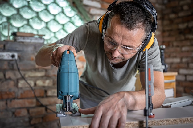 A Caucasian carpenter is concentrating on cutting wood with a jigsaw in his workshop.