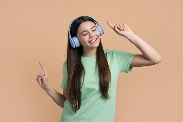 Caucasian cheerful happy smiling teen girl dancing listening to music on blue wireless headphones Lovely adolescent child have fun points fingers while dancing over isolated beige color background