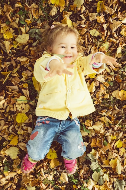 Caucasian child girl dressed in a jacket lies on the ground in autumn leaves.