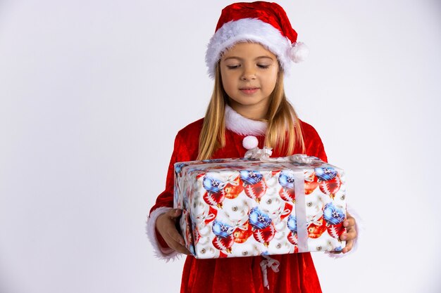 Caucasian child in Santa red hat holding Christmas gift in hand and looking at him. Isolated on