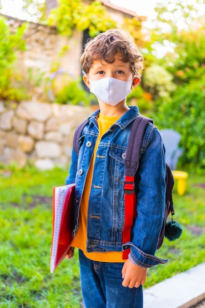 Caucasian child with face mask ready for back to school. New normality, social distance, coronavirus pandemic, covid-19. Jacket, backpack and a red block for notes in hand