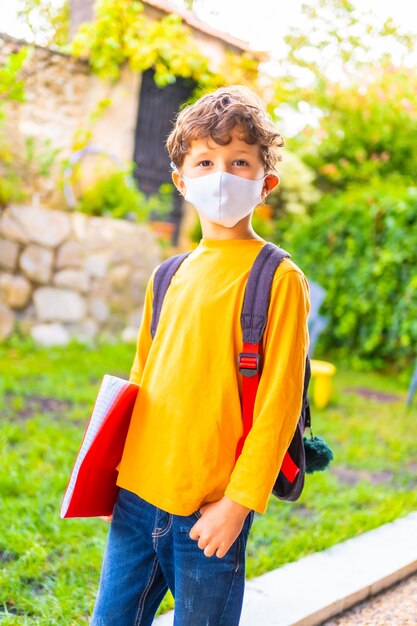 Caucasian child with face mask ready for back to school. New normality, social distance, coronavirus pandemic, covid-19. Orange T-shirt, backpack and a notepad in hand