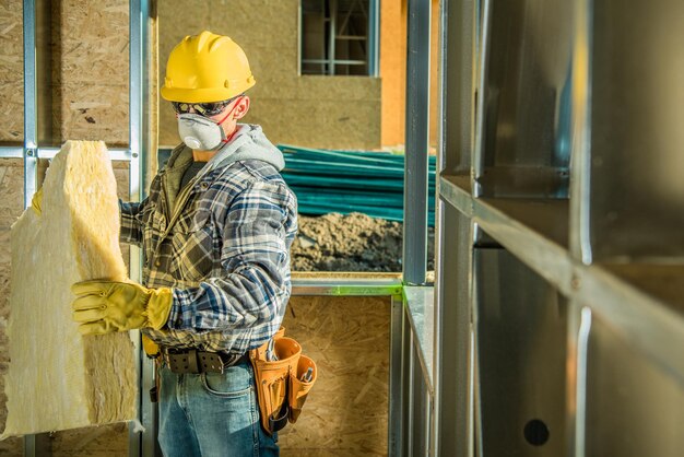 Photo caucasian contractor worker in safe breathing mask and hard hat working inside developed building moving mineral wool insulation construction industry safety