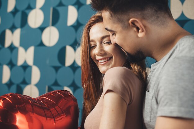 Caucasian couple celebrating valentines day together embracing on a blue wall and holding red air balloons