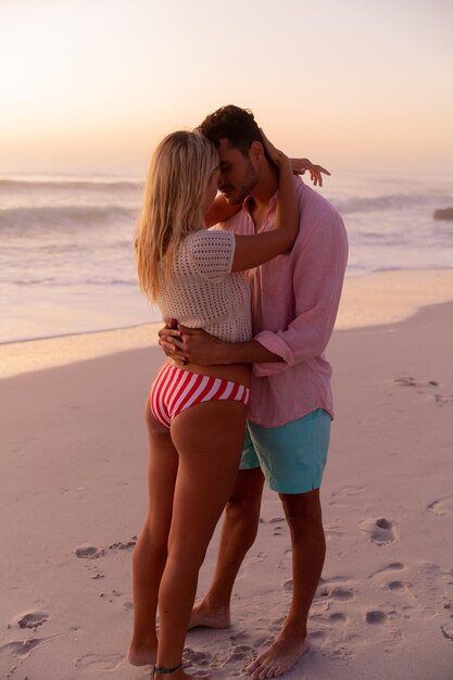 Caucasian couple enjoying time at the beach during a pretty sunset, embracing and kissing with blue sky and sea in the background