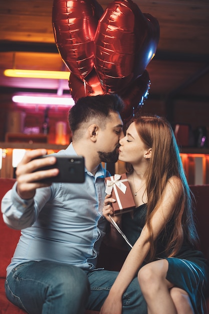 Caucasian couple kissing on a red couch holding air balloons and making a selfie with phone
