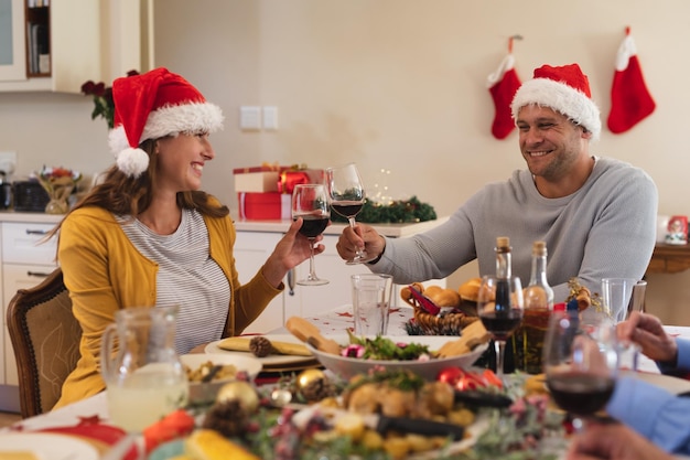 Caucasian couple sitting at table for dinner together, wearing santa hats, holding glasses of wine and making a toast. quality family time christmas celebration.